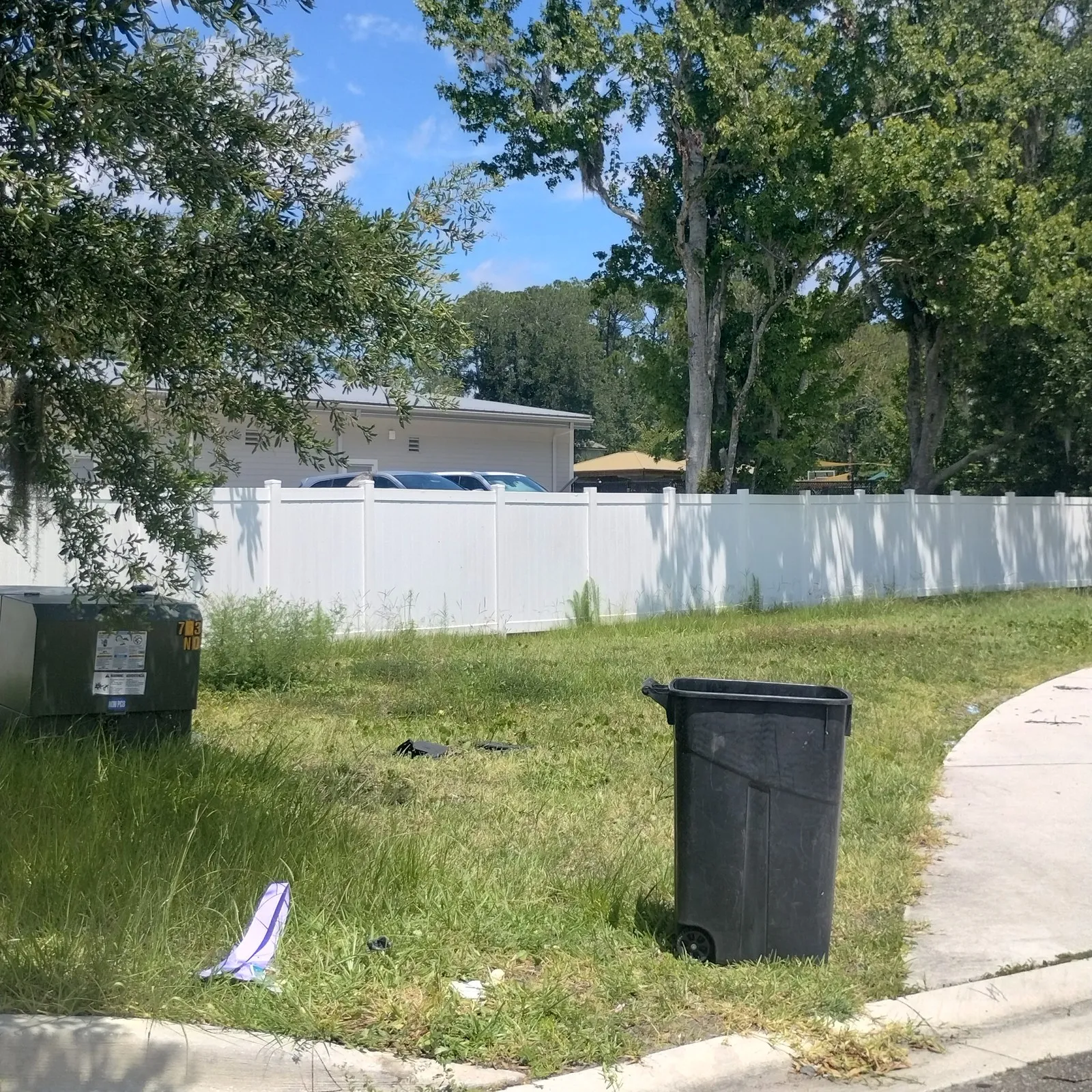 Overgrown common area along a white privacy fence with litter and a trash bin